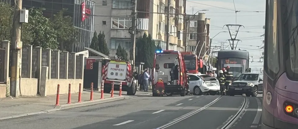 Un vehicul de serviciu de pompieri, o ambulanță și un autoturism alb sunt parcate pe o stradă lângă un tramvai și clădiri urbane.