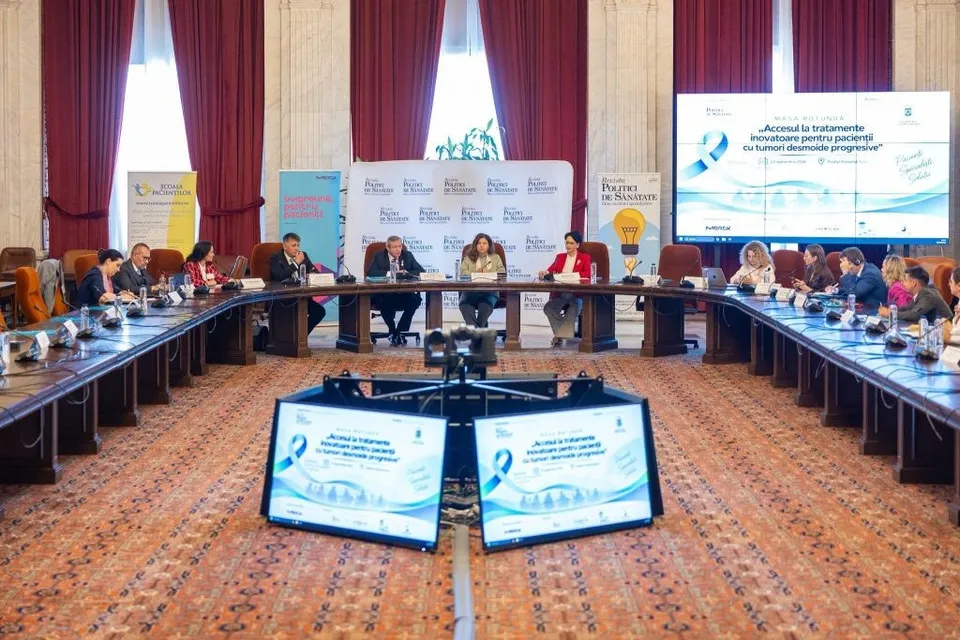 Oamenii stau așezați la mese lungi în jurul unei săli de conferințe, cu ecrane și panouri informative în fundal.