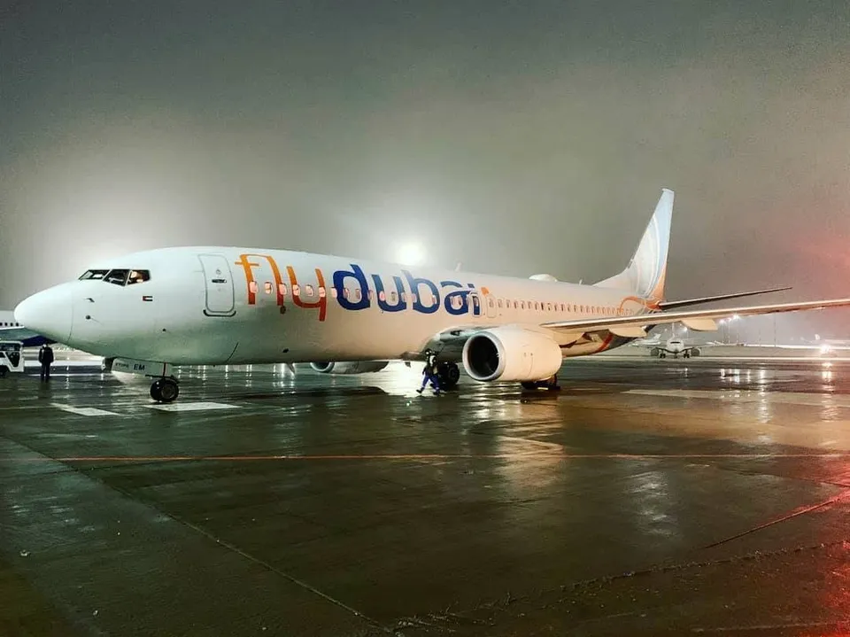 Un avion alb cu inscripția flydubai stă pe o pistă umedă sub un cer nocturn și întunecat.