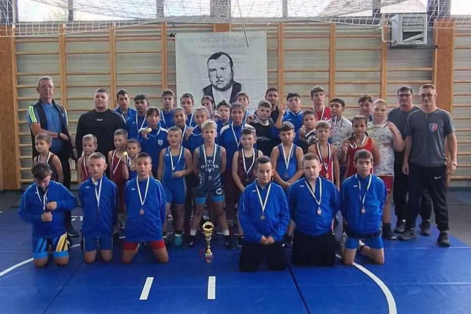 Un grup de sportivi tineri în echipamente sportive albastre și negre pozează pe un covor de wrestling albastru în fața unui poster.