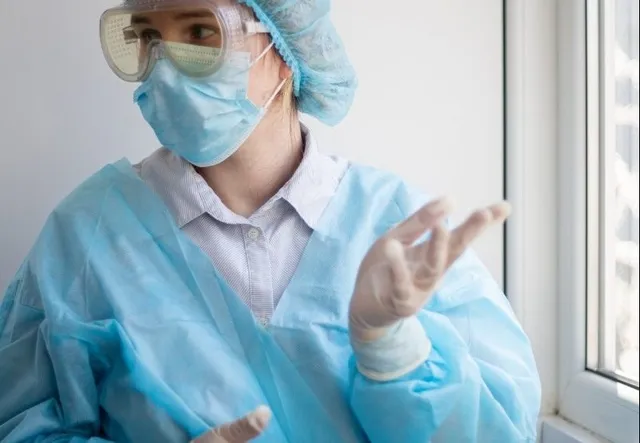 O persoană poartă o mască de protecție albastră, ochelari de protecție, o cască de păr și un halat medical albastru în fața unei ferestre.