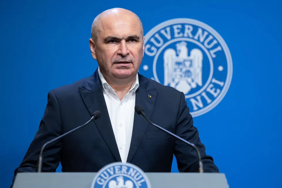 Un bărbat cu părul scurt și costum închis stă în fața unui fundal albastru pe care apare un sigiliu circular cu un heraldic.