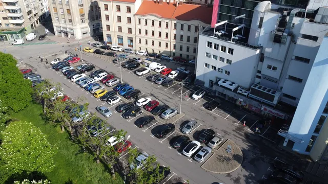 O vedere de sus prezintă o parcare cu numeroase mașini colorate parcate în rânduri între clădiri și un gard de vegetație verde.