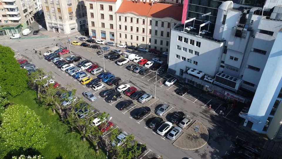 O vedere de sus prezintă o parcare cu numeroase mașini colorate parcate în rânduri între clădiri și un gard de vegetație verde.