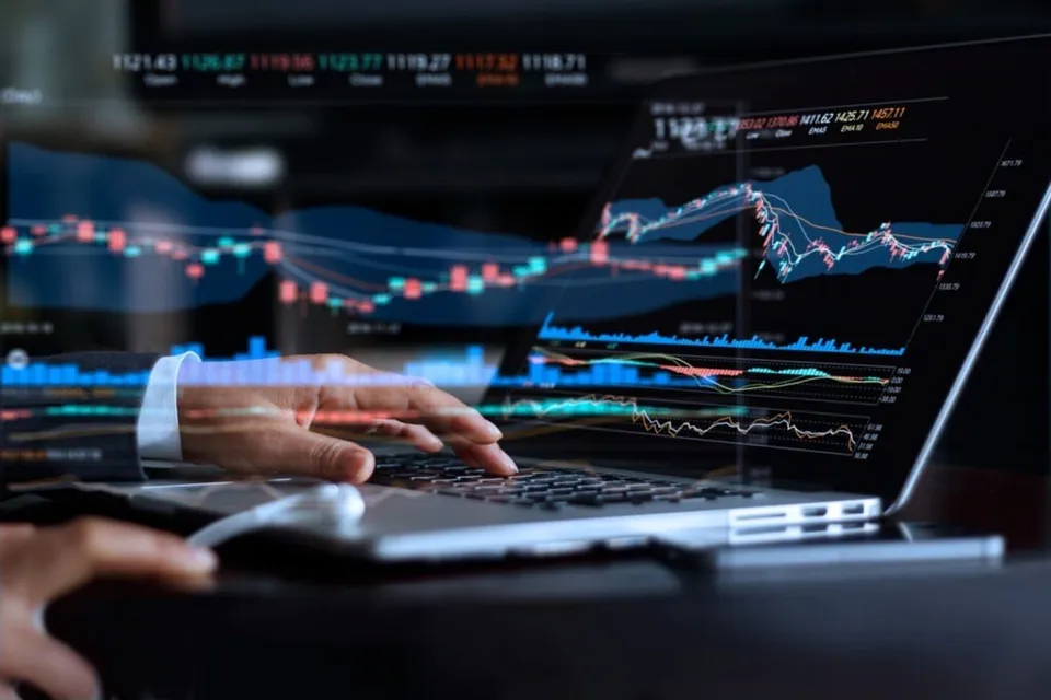 O mână de pe un laptop afișează grafice financiare și indicatori de piață pe un ecran în fundal întunecat.