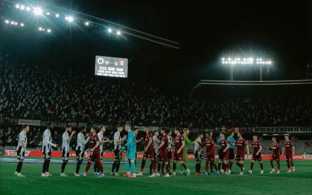 Jucători de fotbal în echipamente de joc stau în rând pe terenul unui stadion umplut de spectatori sub lumini puternice de iluminat.
