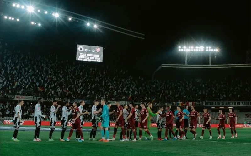 Jucători de fotbal în echipamente de joc stau în rând pe terenul unui stadion umplut de spectatori sub lumini puternice de iluminat.