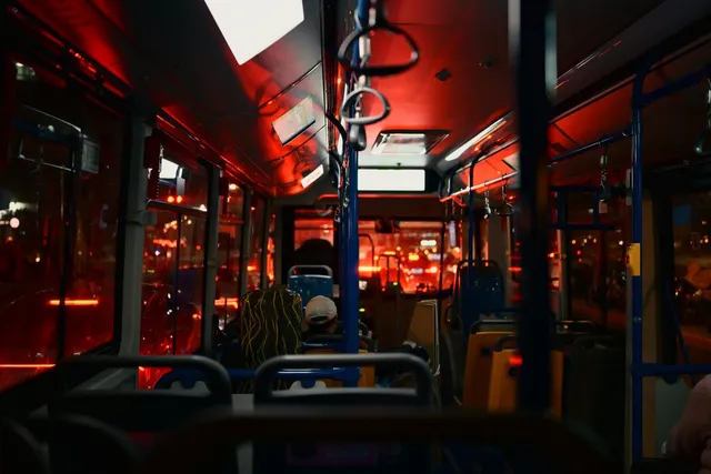 Interiorul unui autobuz iluminat de lumini roșii, cu scaune, mânere de susținere și ferestre prin care se văd lumini de oraș noaptea.