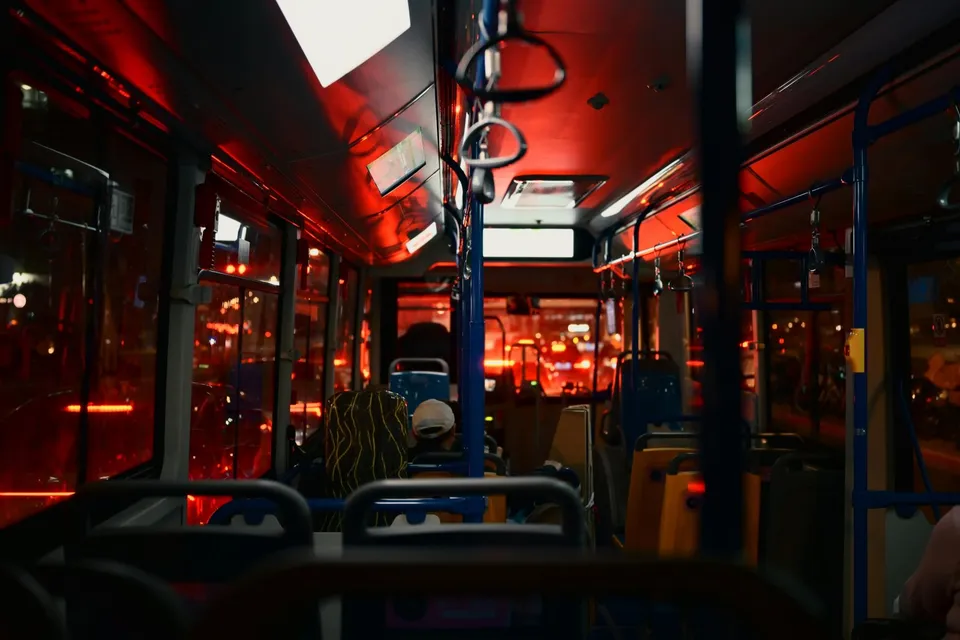 Interiorul unui autobuz iluminat de lumini roșii, cu scaune, mânere de susținere și ferestre prin care se văd lumini de oraș noaptea.
