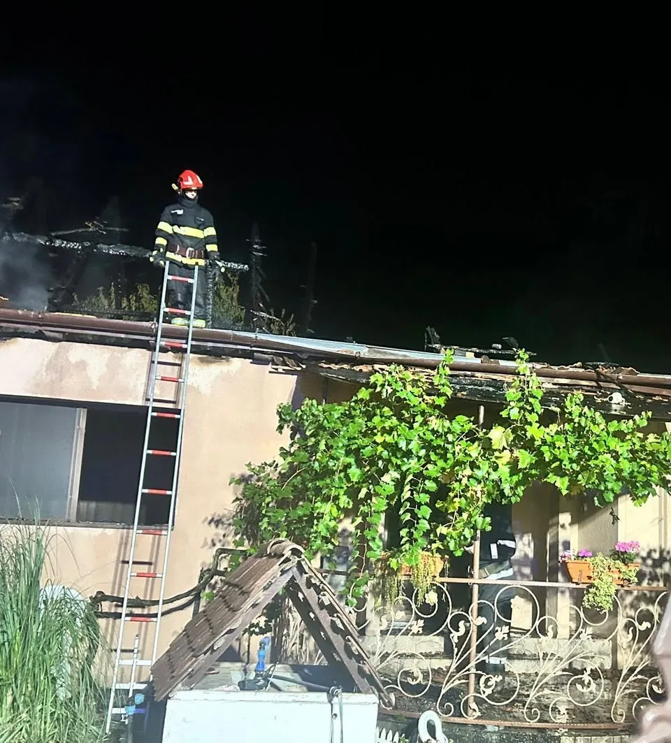 Un pompier cu cască roșie stă pe acoperișul unei clădiri lângă o scară metalică lungă sub un cer de noapte.