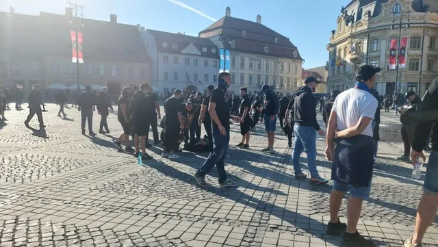 Un grup de persoane merge sau stă pe o piață pavată cu pietriș, în fața unor clădiri urbane sub un cer senin.