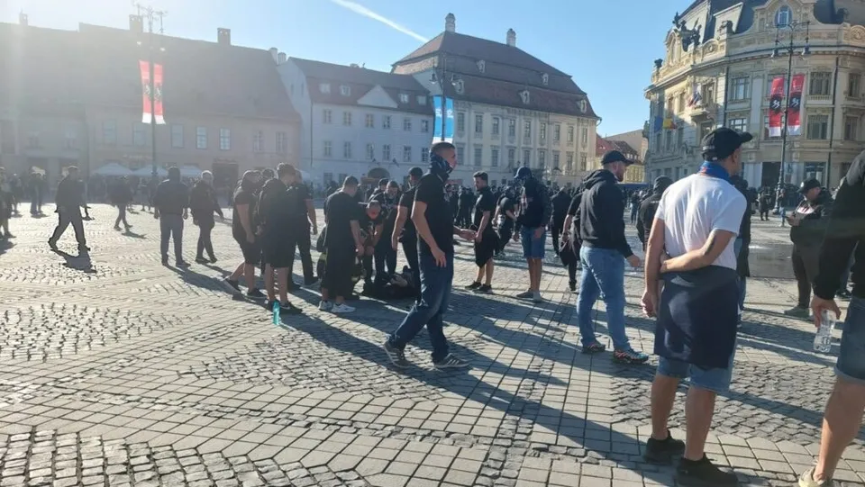 Un grup de persoane merge sau stă pe o piață pavată cu pietriș, în fața unor clădiri urbane sub un cer senin.