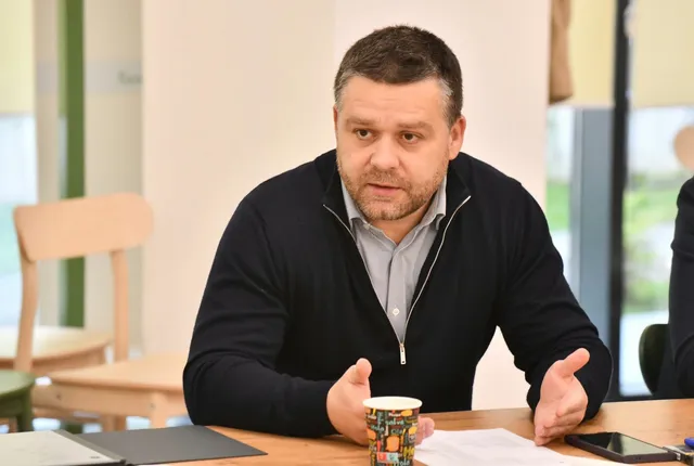 Un bărbat cu părul scurt și cămașă deschisă sub un cardigan închis la culoare stă la o masă de lemn cu mâinile ridicate.