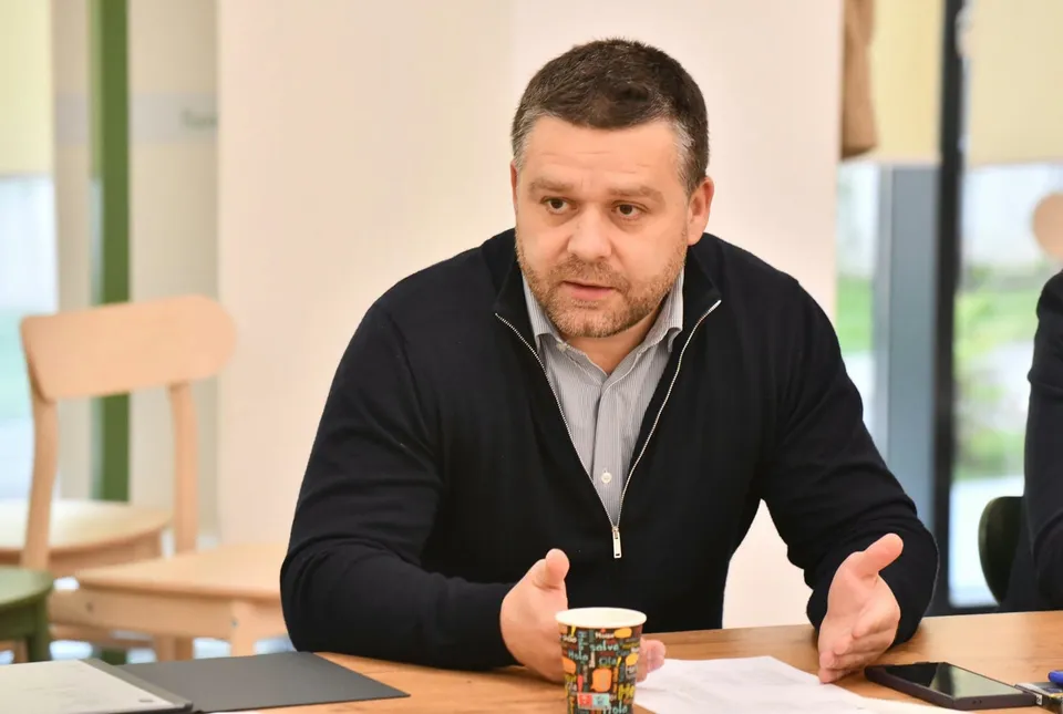 Un bărbat cu părul scurt și cămașă deschisă sub un cardigan închis la culoare stă la o masă de lemn cu mâinile ridicate.