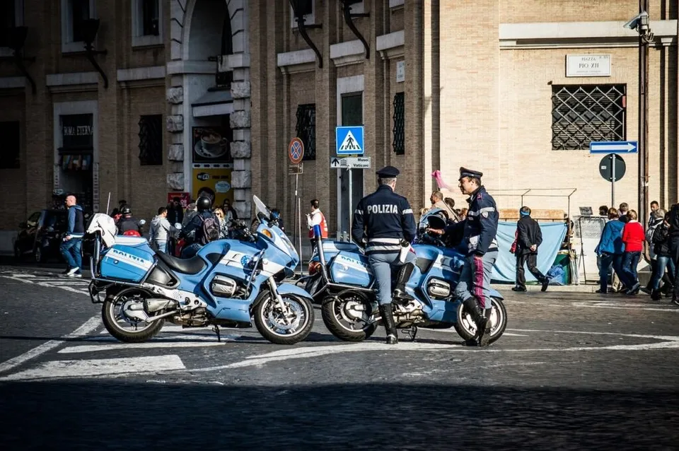 Două motociclete albastre de poliție sunt parcate pe o stradă lângă doi polițiști în uniformă care stau lângă ele.