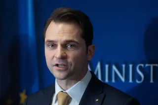 Un bărbat cu părul scurt și deschis la culoare poartă sacou închis, cămașă albă și cravată galbenă, vorbind în fața unui fundal albastru cu text parțial vizibil.