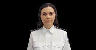 O femeie cu părul brunet poartă o cămașă albă cu epoleți negri și stă în fața unui fundal negru.