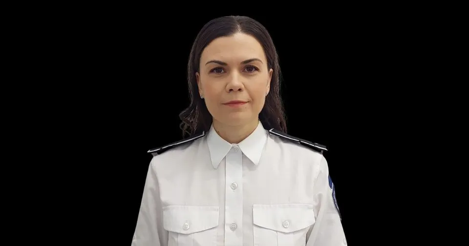 O femeie cu părul brunet poartă o cămașă albă cu epoleți negri și stă în fața unui fundal negru.