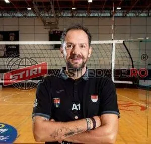 Un bărbat cu barbă și tatuaje pe brațe stă cu brațele încrucișate purtând un tricou polo negru în fața unei săli de sport.