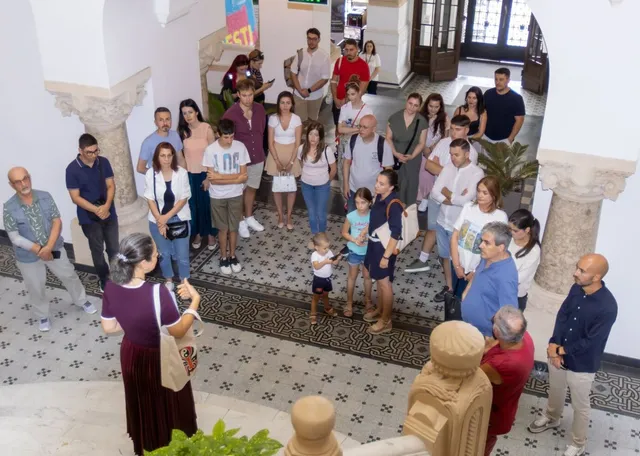 Un grup de oameni stă adunat într-un hol cu podea decorată, ascultând o femeie cu părul scurt și haine închise la culoare.