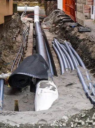 Mai multe conducte de culoare gri și o conductă albă sunt dispuse într-un șanț excavat în pământ.