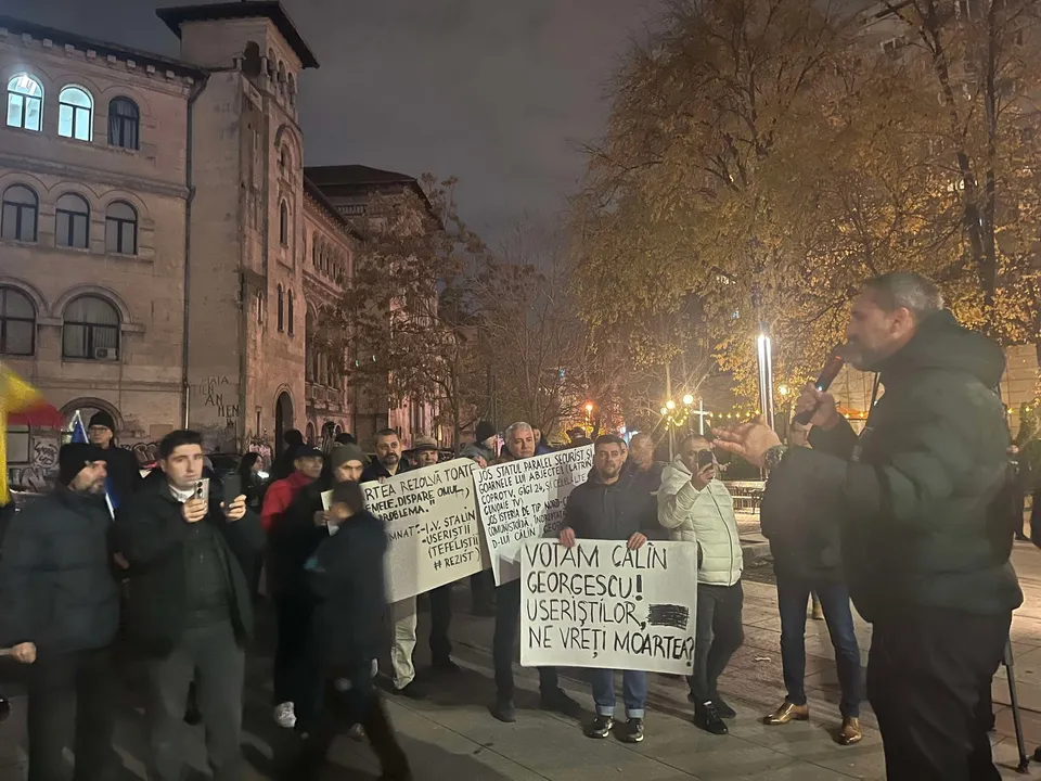 Un grup de persoane stă într-o piață noaptea, purtând panouri cu mesaje scrise și ascultând un bărbat care vorbește într-un microfon.