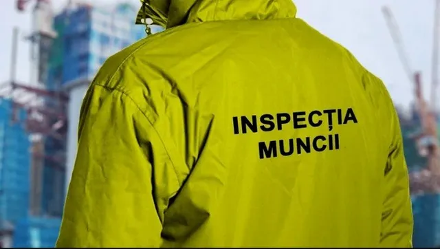 Un bărbat poartă o jachetă galbenă pe care este scris textul negru INSPECTIA MUNCII, având în fundal o clădire în construcție.