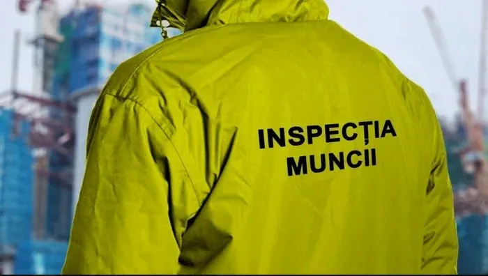 Un bărbat poartă o jachetă galbenă pe care este scris textul negru INSPECTIA MUNCII, având în fundal o clădire în construcție.