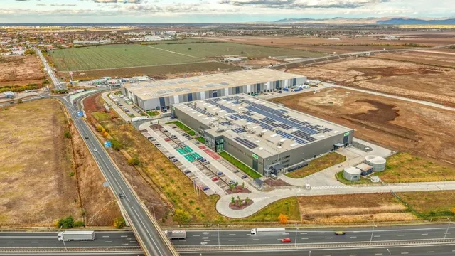O vedere aeriană prezintă o clădire industrială mare cu un parc de parcare și o autostradă în prim-plan, înconjurată de câmpuri deschise.