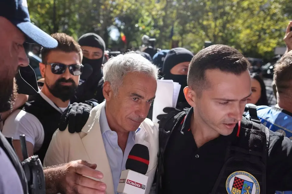 Un bărbat cu părul cărunt și sacou deschis este înconjurat de mai mulți bărbați, inclusiv unul cu ochelari de soare și unul în vestă tactică.