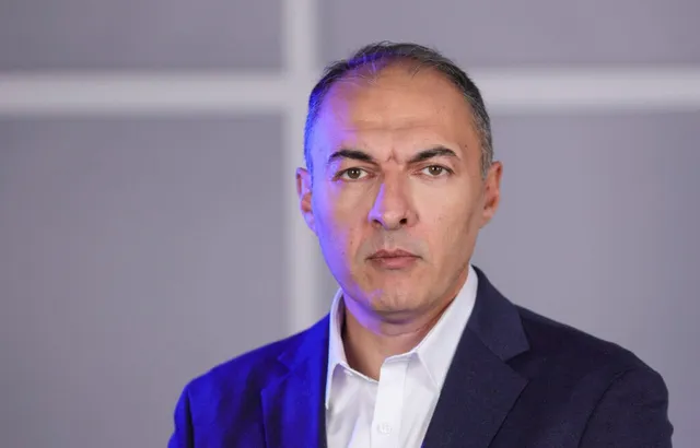 Un bărbat cu părul scurt și cămașă albă poartă un sacou închis la culoare în fața unui fundal gri cu linii albe.