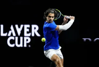Un jucător de tenis cu tricou albastru și pantaloni albi lovește o minge galbenă cu o rachetă pe un teren de tenis.