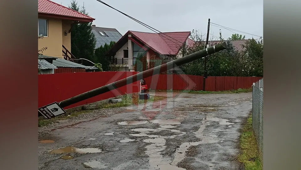 Un stâlp înclinat cu cabluri întinse se află deasupra unui drum de pietriță și asfalt cu bălți, lângă un gard roșu și case.
