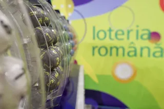 Numerele sunt scrise pe bilete rotunde de culoare gri aflate într-un aparat de extragere transparent, lângă un panou colorat cu text.