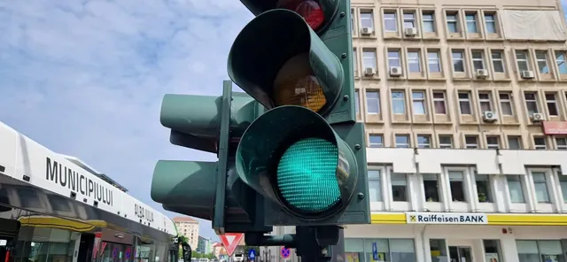 Un semafor cu lumina verde aprinsă este montat pe un stâlp, având în fundal o clădire cu ferestre și un cer înnorat.