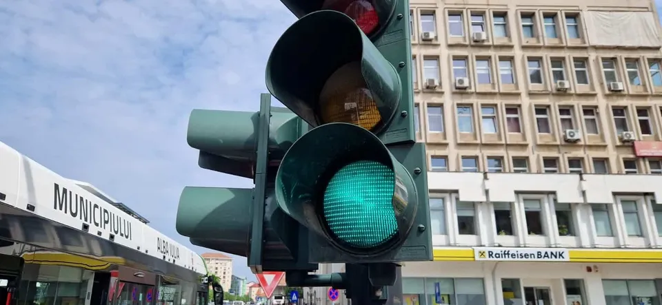 Un semafor cu lumina verde aprinsă este montat pe un stâlp, având în fundal o clădire cu ferestre și un cer înnorat.