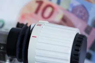Un robinet termostatic alb-negru ocupă prim-planul, cu o bancnotă roz, estompată, în fundal.