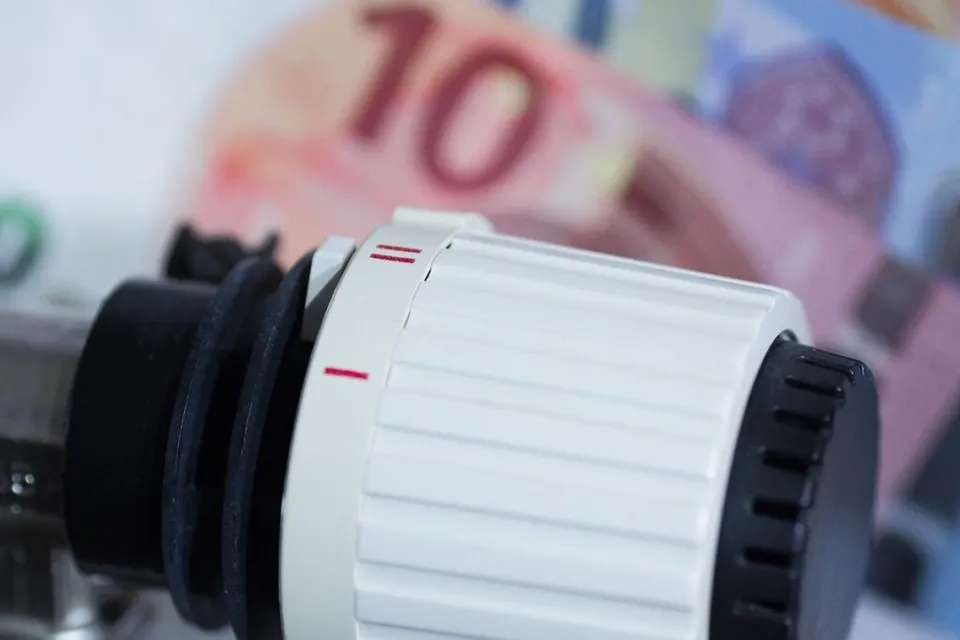 Un robinet termostatic alb-negru ocupă prim-planul, cu o bancnotă roz, estompată, în fundal.