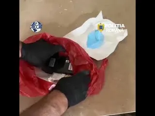 Mâini cu mănuși negre țin o pungă de plastic roșie deschisă pe o suprafață plană lângă un obiect alb și albastru.
