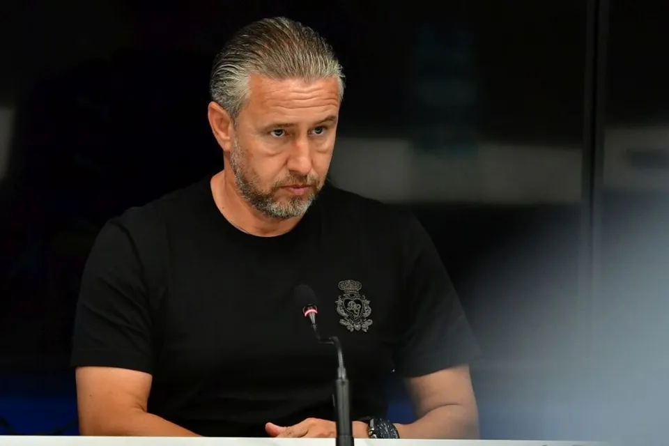 Un bărbat cu părul cărunt și barbă scurtă poartă un tricou negru cu un emblema pe piept, stând în fața unui microfon.