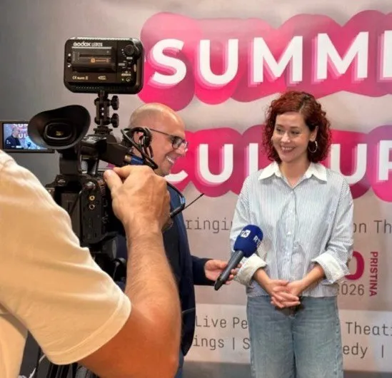 O femeie zâmbește în timp ce este filmată de o cameră video în fața unui panou roz și alb.
