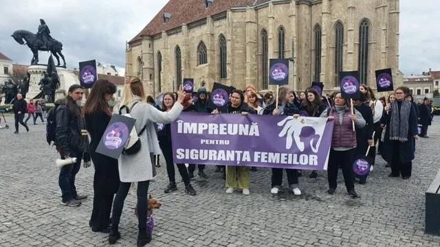Un grup de persoane poartă un banner violet cu text alb și diverse pancarte mici în fața unei clădiri mari de piatră.