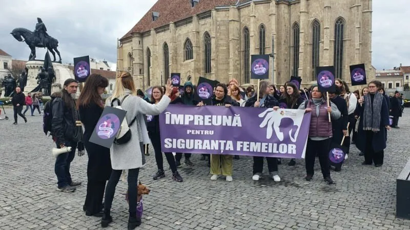 Un grup de persoane poartă un banner violet cu text alb și diverse pancarte mici în fața unei clădiri mari de piatră.