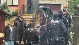 O persoană în haine civile este însoțită de mai mulți polițiști în echipament tactic cu inscripția 'POLIȚIA' pe spate, în fața unei locuințe.