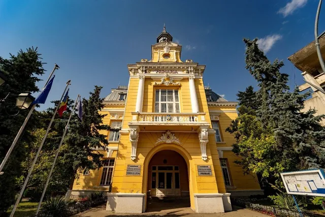 O clădire galbenă cu arhitectură clasică, având ferestre mari, un balcon alb și steaguri montate pe stâlpi în partea stângă.