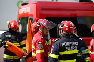 Patru persoane purtând uniforme de serviciu și căști de protecție roșii stau lângă un vehicul de culoare roșie.