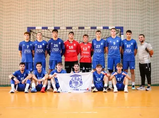 Un grup de jucători de handbal în echipamente albastre și roșii pozează în fața unei ținte de handbal pe un teren de sport.