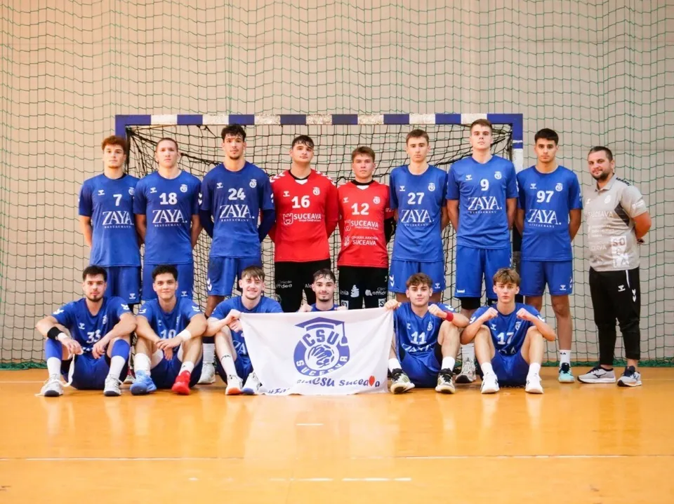 Un grup de jucători de handbal în echipamente albastre și roșii pozează în fața unei ținte de handbal pe un teren de sport.