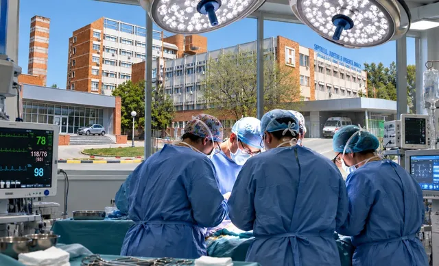 Patru chirurgi în halate albastre operează sub lumini chirurgicale, cu un monitor medical afișând date vitale. În fundal, o clădire mare, posibil un spital, și o ambulanță.