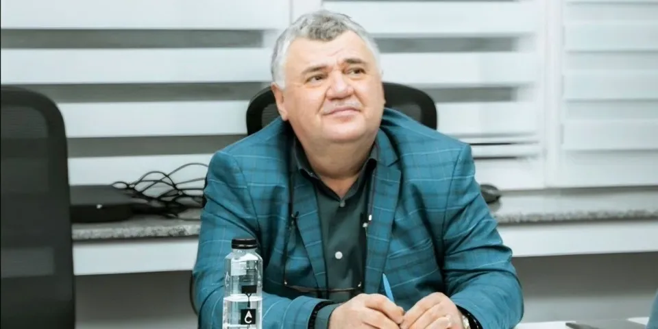 Un bărbat în vârstă, cu păr gri, îmbrăcat într-o jachetă albastră și o camășie neagră, este așezat la o masă, ținând un pix în mână.